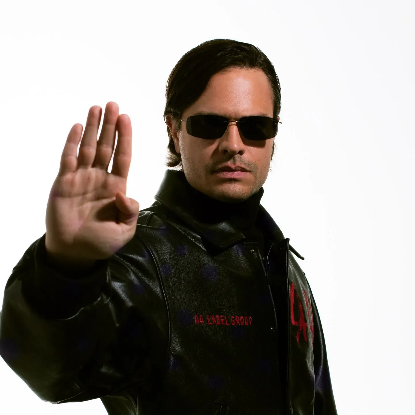 Retrato promocional de un DJ con gafas oscuras, chaqueta de cuero negra y la mano levantada sobre fondo blanco, imagen de estética electrónica.