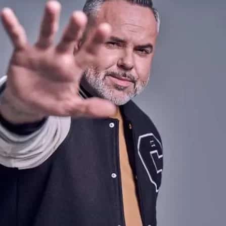 Retrato promocional de un DJ con chaqueta universitaria, barba canosa y la mano extendida hacia la cámara sobre un fondo gris neutro.