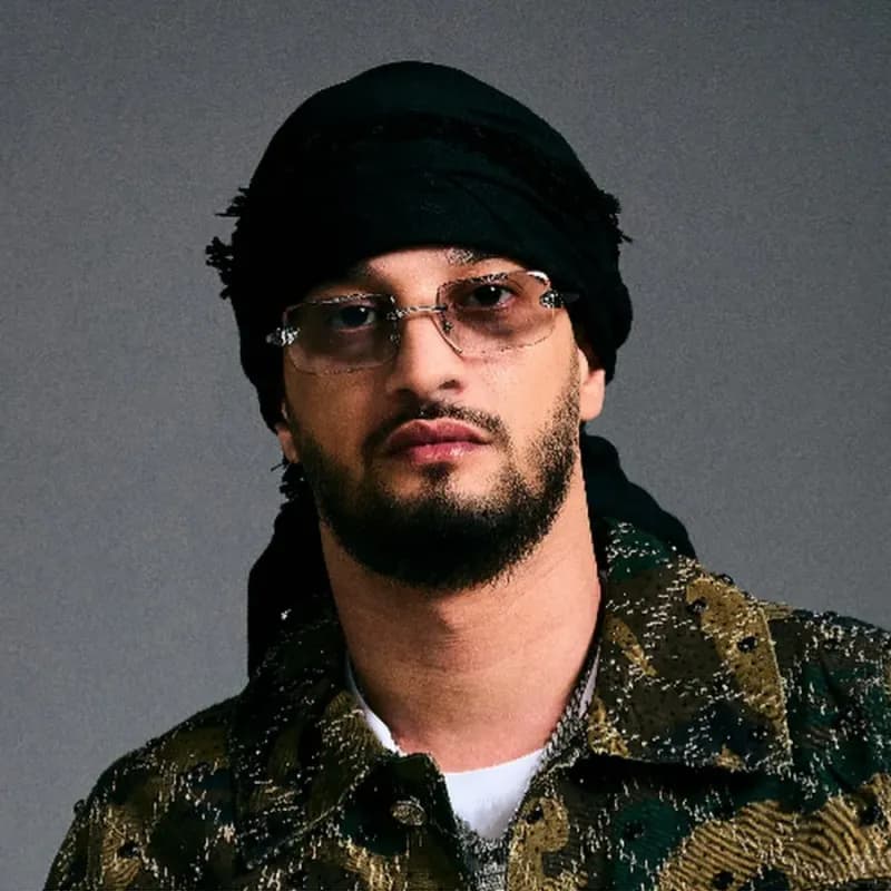 Retrato de Soolking con gafas y pañuelo negro sobre fondo gris, imagen del cantante argelino de rap francófono, raï y música urbana.