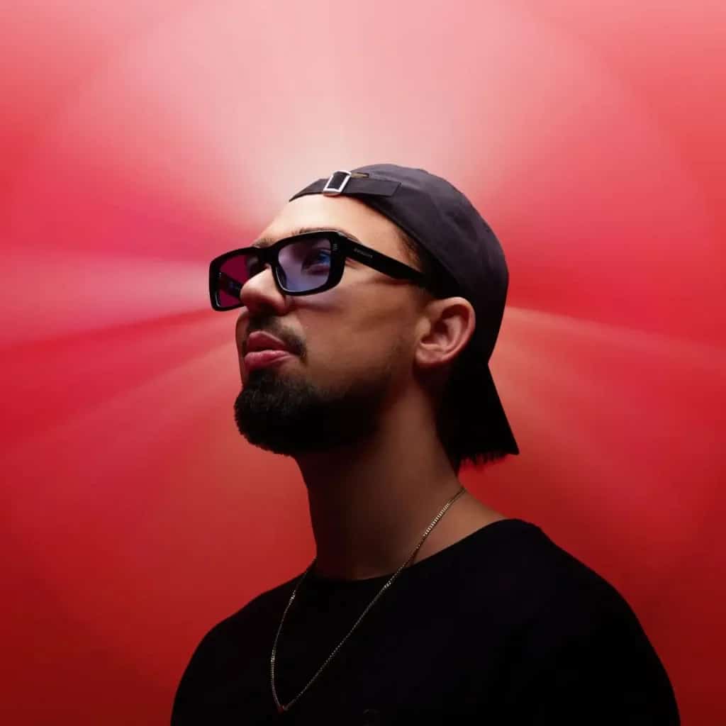 Portrait of Sonny Fodera wearing dark sunglasses and a black cap against a red background, image of the Australian melodic and vocal house DJ.