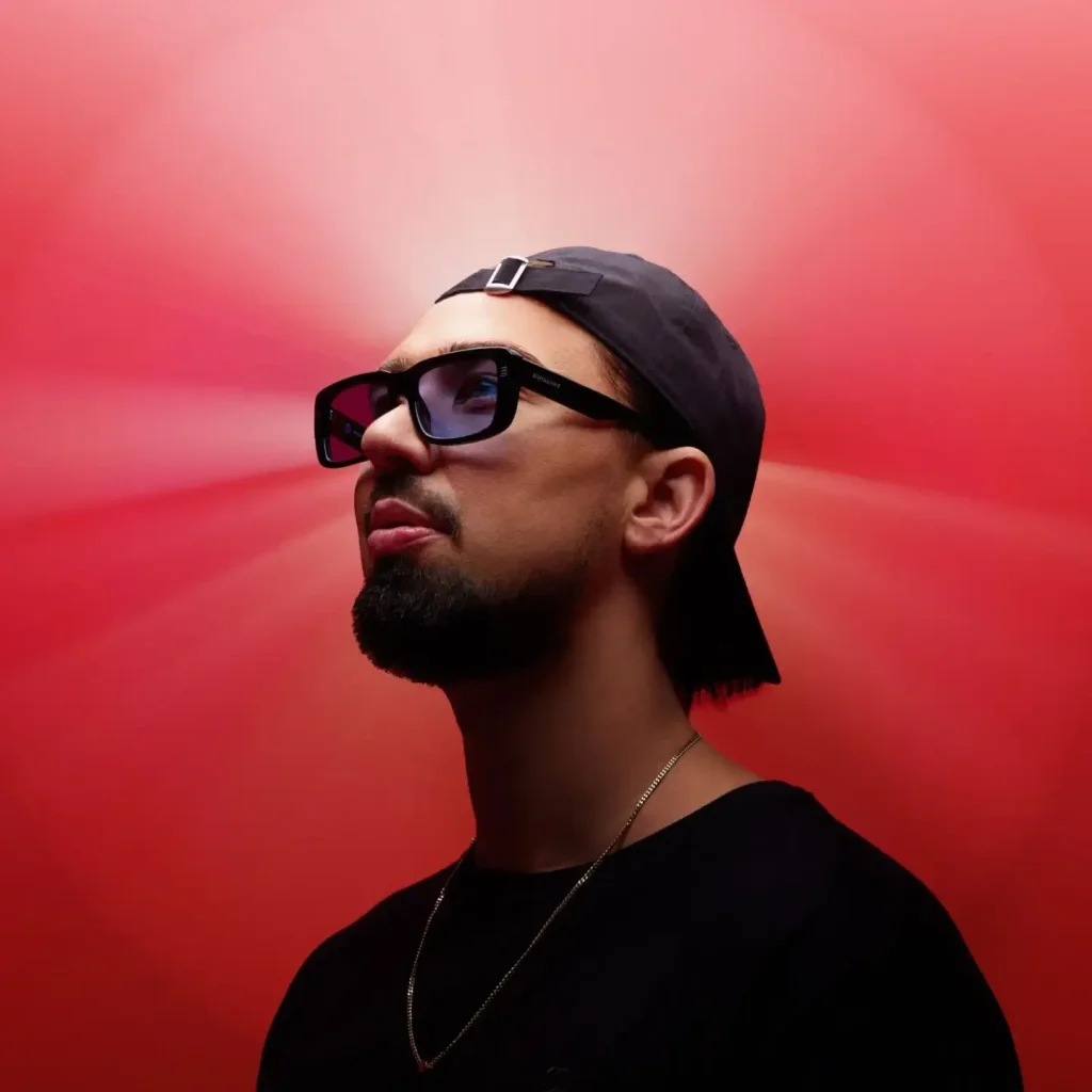 Portrait of Sonny Fodera wearing dark sunglasses and a black cap against a red background, image of the Australian melodic and vocal house DJ.