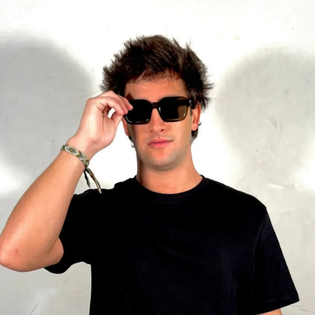 DJ joven con gafas de sol y camiseta rosa de espaldas sobre fondo oscuro, imagen promocional de música house y tech-house.