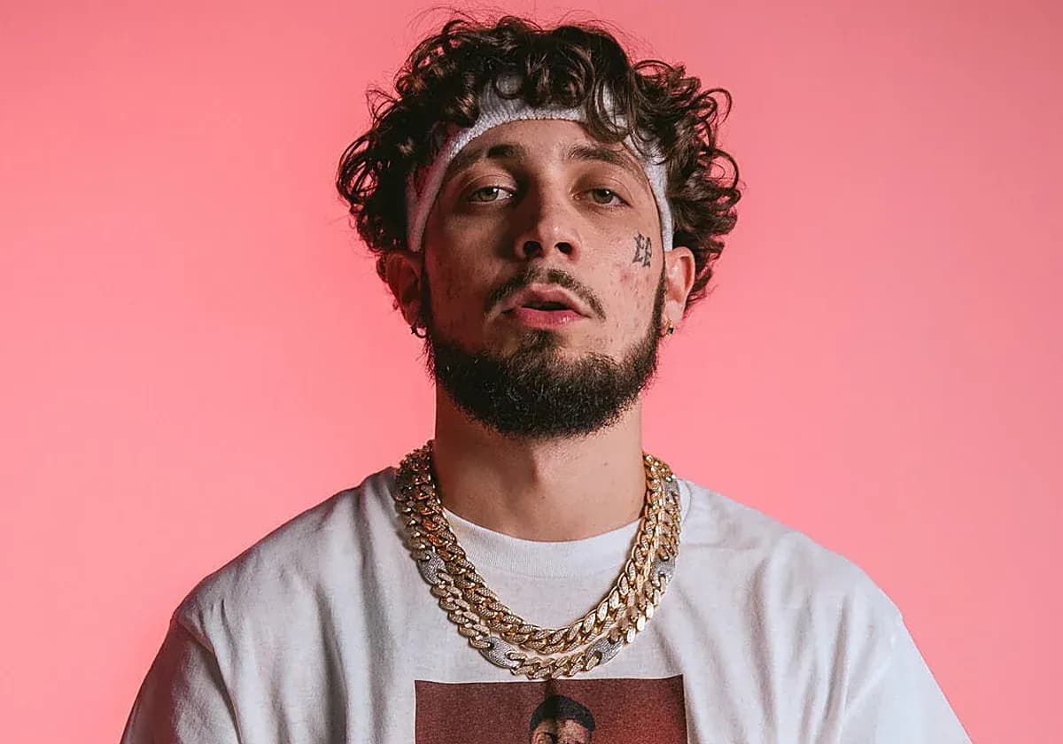 Retrato frontal de JC Reyes con cinta blanca, cadenas doradas y camiseta blanca sobre fondo rosa, imagen del cantante y rapero urbano sevillano.