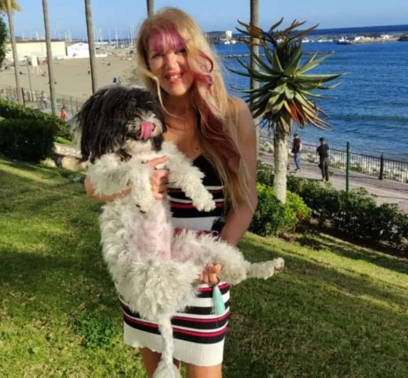 Loulou sonriendo mientras sostiene un perro blanco y negro frente al mar en Marbella, con palmeras y paseo marítimo de fondo. Imagen representando la comunidad social “Los Amigos de Lou Lou”.