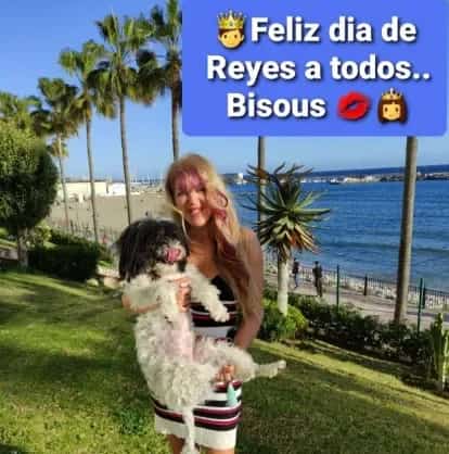 Lou Lou sonriendo mientras sostiene un perro blanco y negro frente al mar en Marbella, con palmeras y paseo marítimo de fondo. Imagen representando la comunidad social “Los Amigos de Lou Lou”.