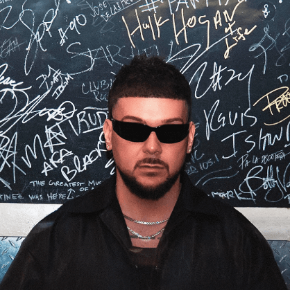 Retrato del DJ madrileño José De La Heras con gafas de sol negras y cadenas plateadas, frente a una pared llena de firmas; imagen promocional de reggaetón y latin house.