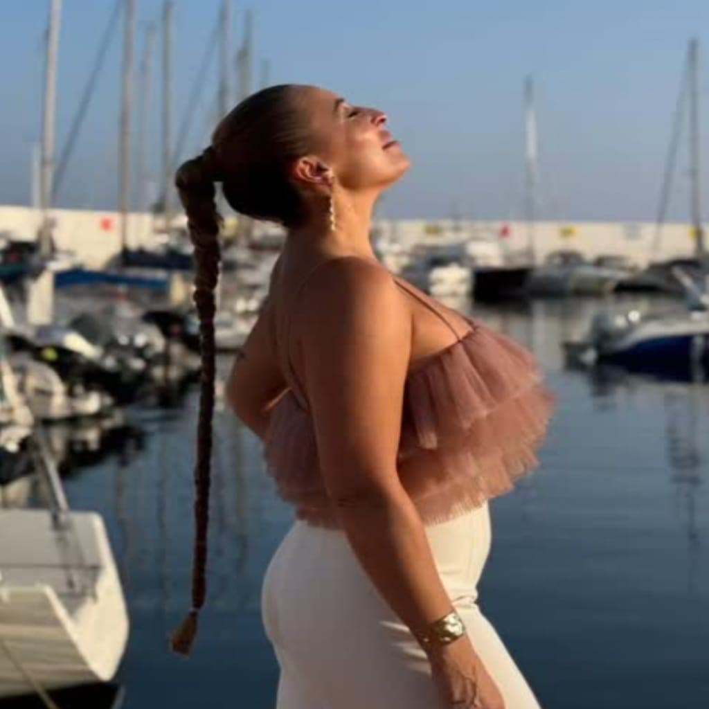 Perfil de Davinia Escalona mirando al cielo sobre el muelle de Marbella; luce top rosa con volantes, pantalón blanco y larga trenza mientras los mástiles de los barcos se recortan al fondo.