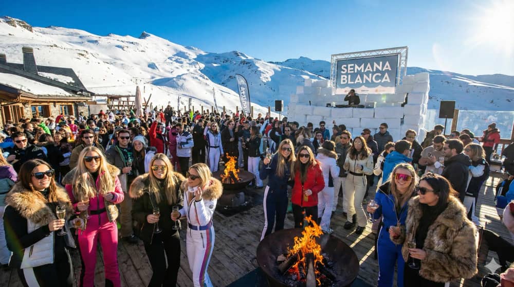 Una fotografía de estilo de vida dinámica y llena de energía de una exclusiva fiesta après-ski en una terraza soleada en Sierra Nevada, España, durante la Semana Blanca. Una multitud de jóvenes con estilo, vestidos con ropa de invierno de diseño y gafas de sol, brindan con bebidas y bailan cerca de hogueras al aire libre. Una cabina de DJ está rodeada de nieve. La luz solar intensa y brillante se refleja en la nieve blanca bajo un cielo azul profundo. El ambiente es de escape alegre y celebración de lujo. Cinematográfica, detallada.
