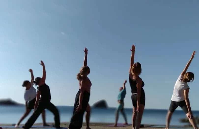 Grupo de personas practicando yoga en la playa al amanecer con los brazos elevados, el mar de fondo en la costa de Marbella.