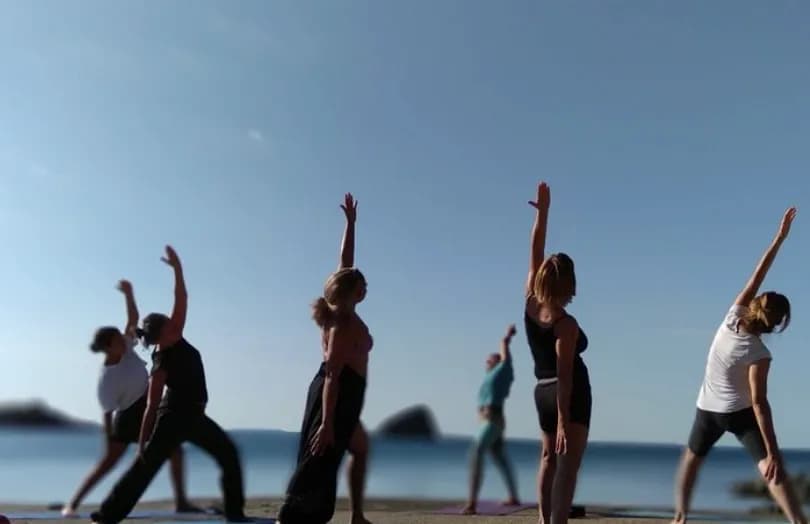 Grupo de personas practicando yoga en la playa al amanecer con los brazos elevados, el mar de fondo en la costa de Marbella.