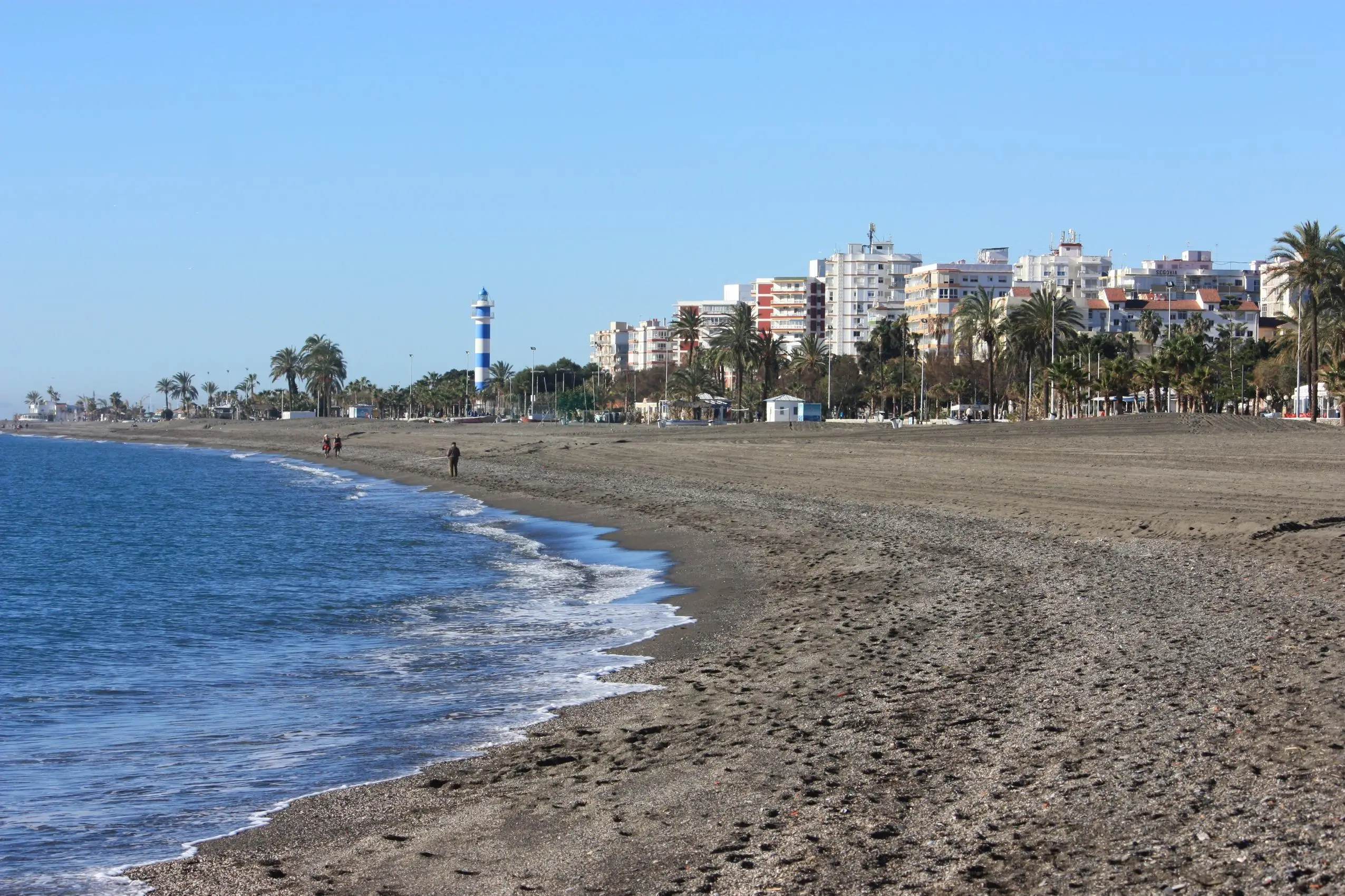 Playa de Torre del Mar con el faro azul y blanco, palmeras y edificios junto a la costa.