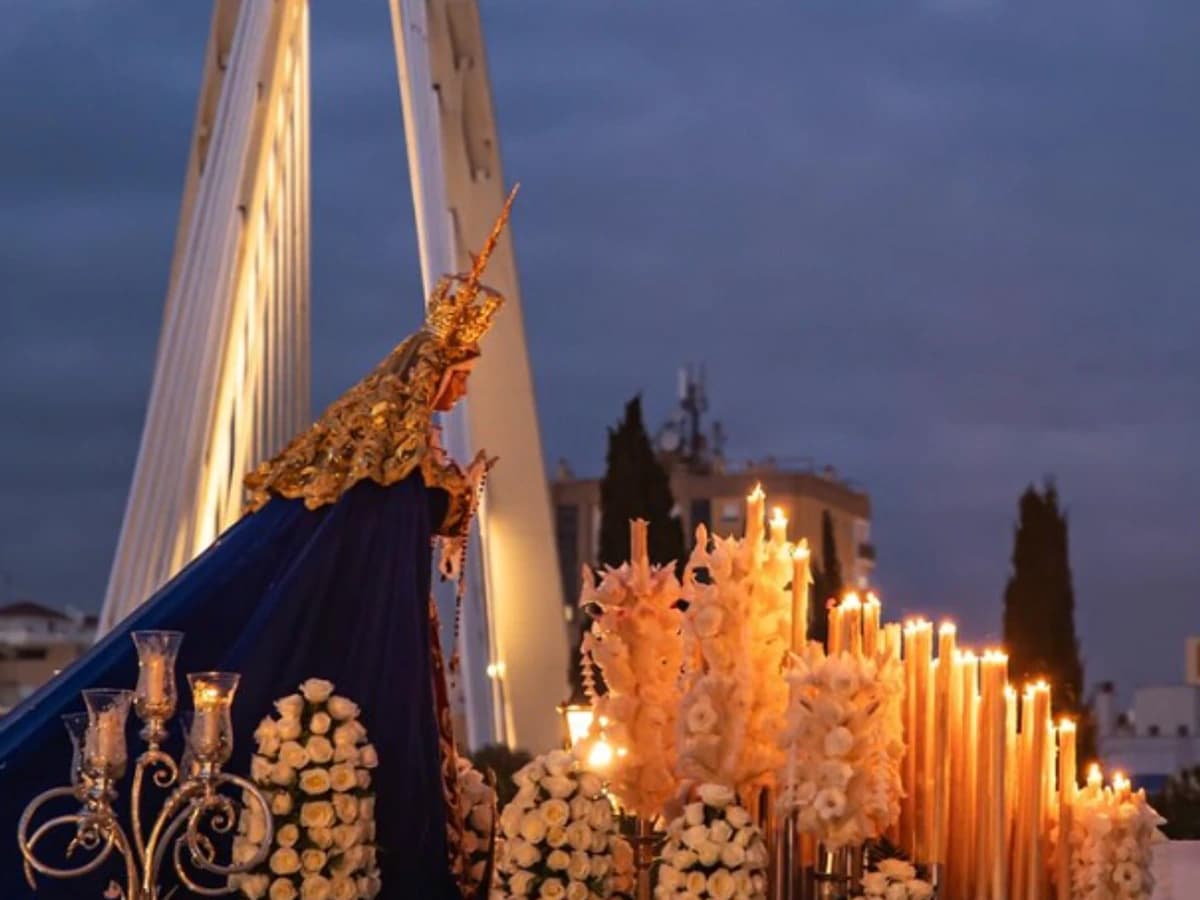 Procesión de Nuestra Señora de la Amargura en Marbella durante el Sábado de Pasión, con el trono de la Virgen iluminado por cirios y rodeado de flores blancas al atardecer, con el puente del arco al fondo.