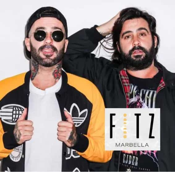 Les Castizos en FITZ Marbella posando frente a un fondo blanco; ambos llevan chaquetas urbanas, uno con gorra hacia atrás, gafas de sol y tatuajes visibles, y el otro con barba y camiseta con diseño gráfico, imagen promocional del evento de Les Castizos en FITZ Marbella.