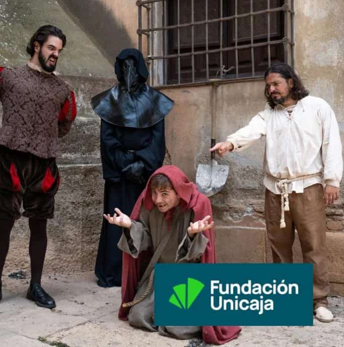 Actors from the play The Plague in 17th-Century Malaga performing a historical scene outdoors, with one character kneeling in a red hooded cloak, another pointing at him with a shovel, a third in period costume and a sinister plague doctor in black mask and cape, in front of an old museum wall.