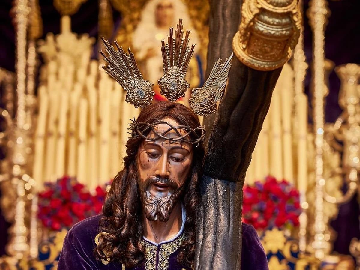 Imagen de Nuestro Padre Jesús Nazareno en Marbella, con túnica morada bordada en oro y la cruz al hombro, en una escena de Semana Santa con fondo de cirios y elementos dorados del trono.