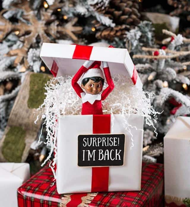 Christmas elf doll popping out of a white gift box with a red ribbon and the sign “Surprise! I’m back”, in front of a snowy Christmas tree, image used for Marbella Creative Kids’ «Elf Is Back!» kids party.