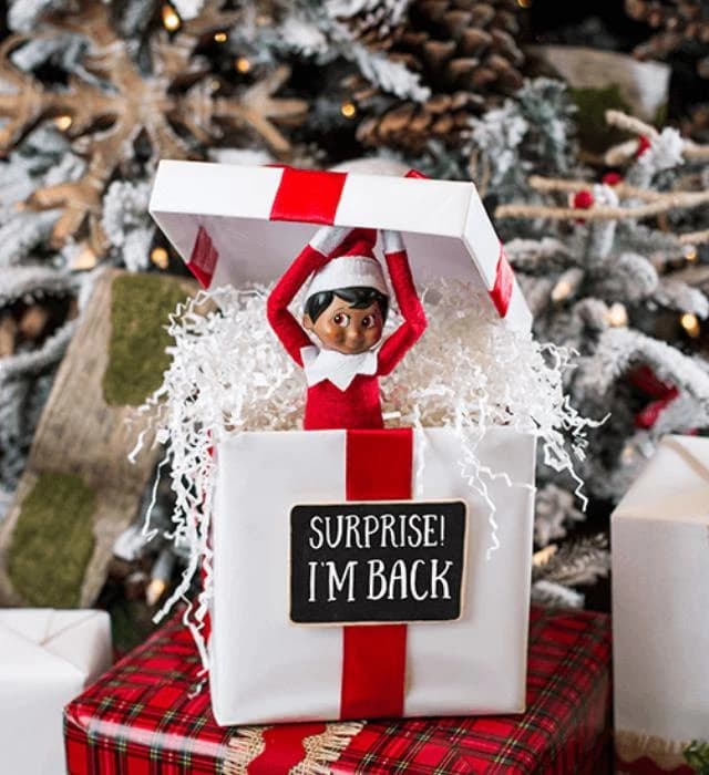 Christmas elf doll popping out of a white gift box with a red ribbon and the sign “Surprise! I’m back”, in front of a snowy Christmas tree, image used for Marbella Creative Kids’ «Elf Is Back!» kids party.