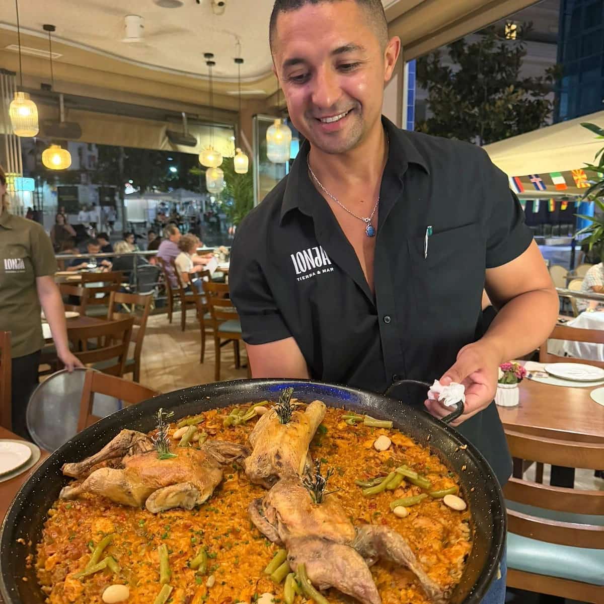 Camarero de La Lonja Marbella mostrando una gran paellera de arroz de carne dentro del restaurante, con el logo del local visible en el uniforme.