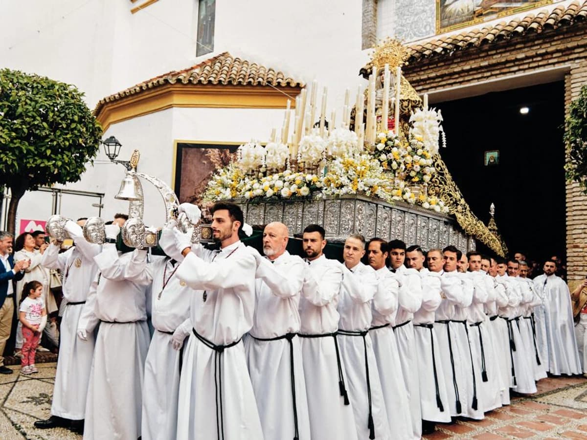 Trono de María Santísima de la Paz y Esperanza de la Cofradía de la Pollinica durante el Domingo de Ramos en Marbella, con portadores vestidos de blanco saliendo del templo entre flores blancas y amarillas.