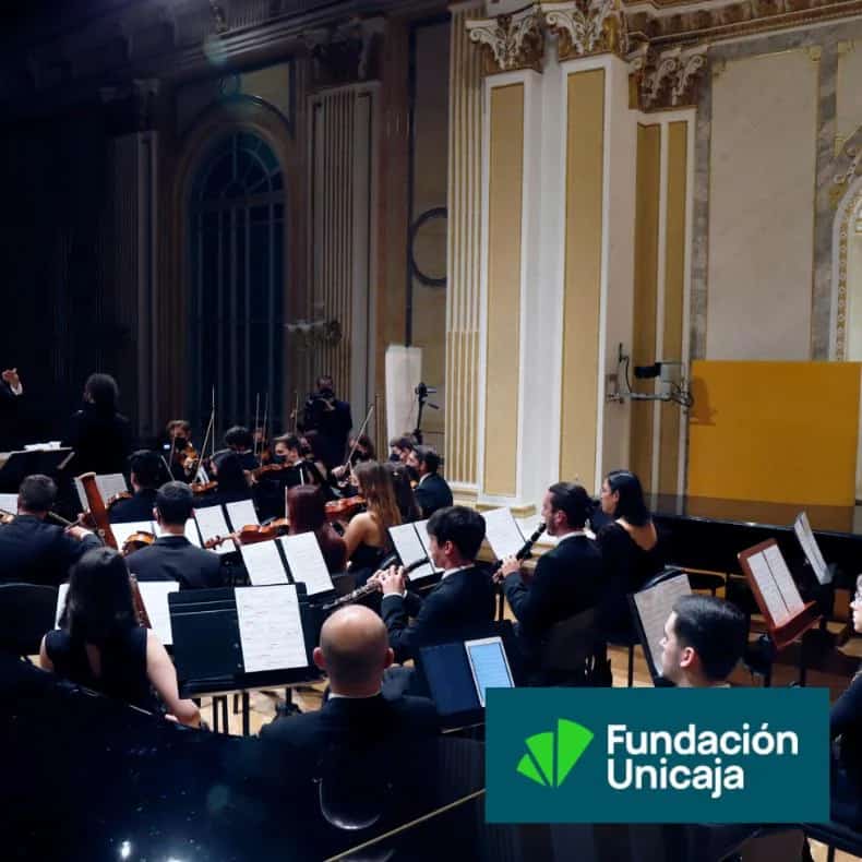 Orquesta sinfónica actuando en directo en la Sala Fundación Unicaja María Cristina de Málaga, con músicos vestidos de gala interpretando sus partituras bajo la dirección del maestro, en un escenario clásico con columnas doradas y decoración histórica.