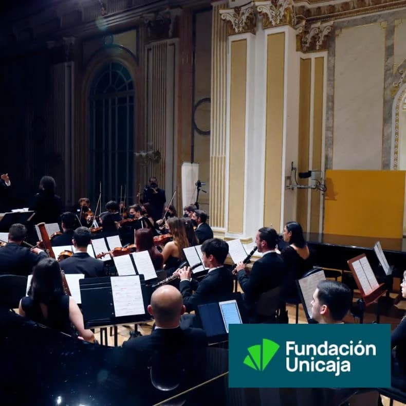 Symphony orchestra performing live at the Unicaja Concert Hall María Cristina in Malaga, with formally dressed musicians playing from their scores under the conductor’s direction, set against a classical stage with golden columns and historic decor.