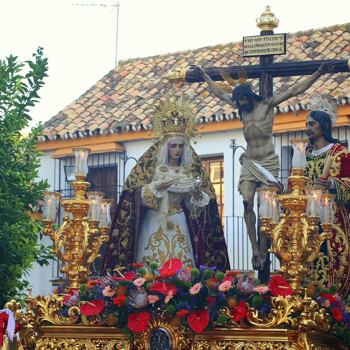 Trono del Cristo del Amor en Marbella durante la Semana Santa, con el Crucificado junto a María Santísima de la Caridad y San Juan Evangelista sobre un paso dorado con flores y candelabros en el centro histórico.