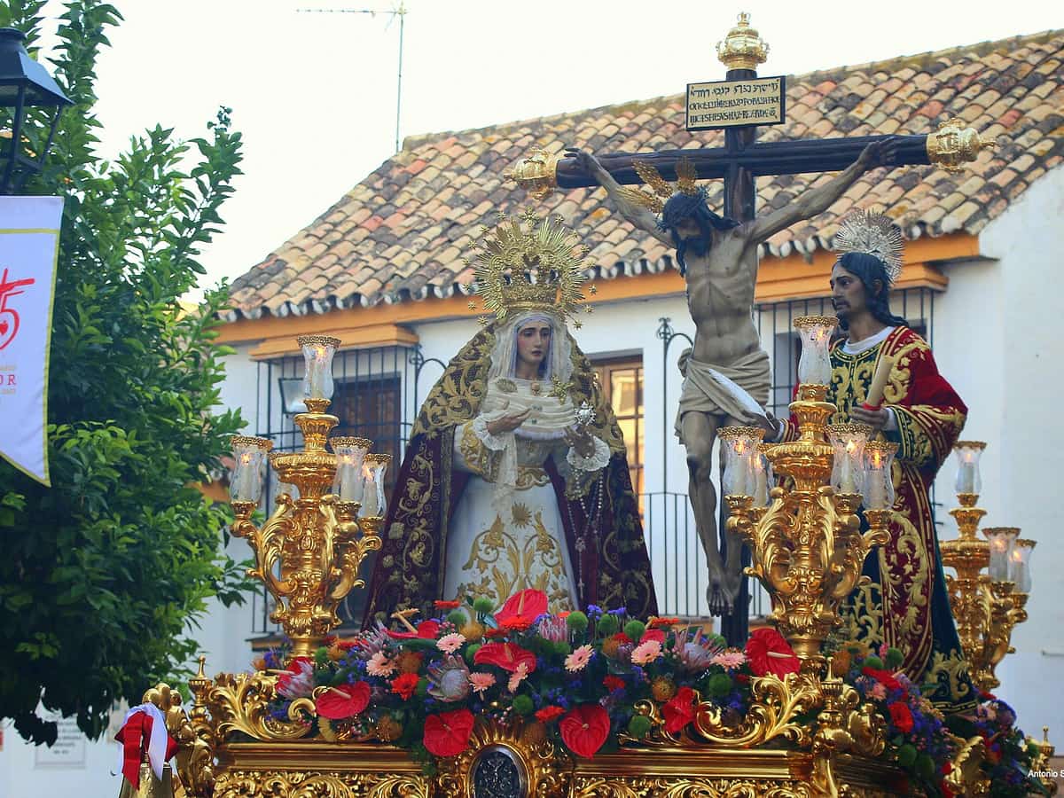 Trono del Cristo del Amor en Marbella durante la Semana Santa, con el Crucificado junto a María Santísima de la Caridad y San Juan Evangelista sobre un paso dorado con flores y candelabros en el centro histórico.