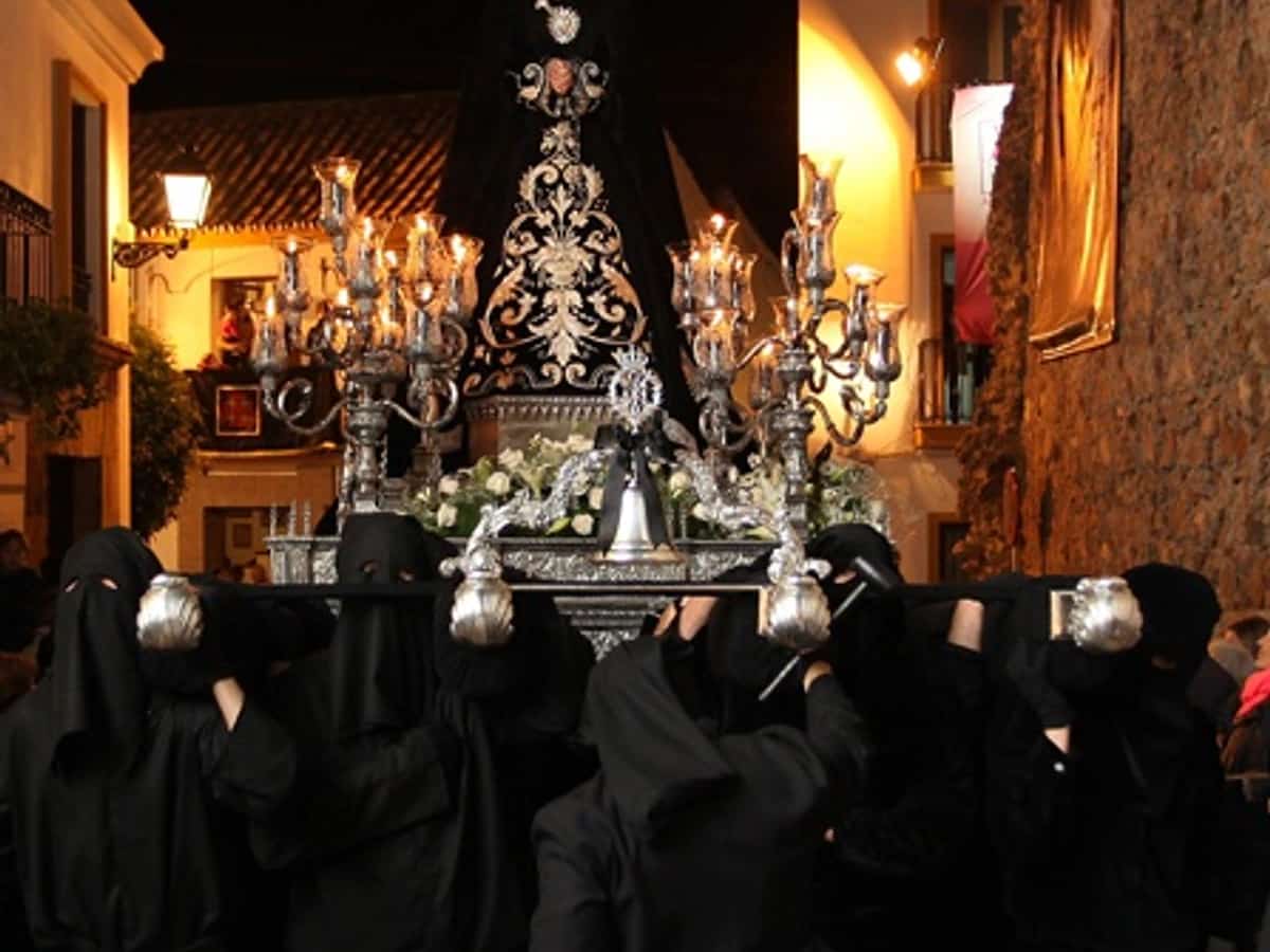 Procesión de Nuestra Señora de la Soledad en Marbella durante el Viernes Santo, con el trono de la Virgen avanzando de noche entre nazarenos de negro, cirios y público en una estrecha calle del centro histórico.