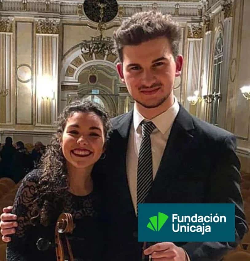 Nazariy Annyuk (violín) y Beatriz Claudio Martínez (violonchelo) uno con violín y otra con violonchelo, posan sonrientes vestidos de concierto en el interior de la Sala Fundación Unicaja María Cristina de Málaga, con las butacas y la decoración clásica del auditorio al fondo.