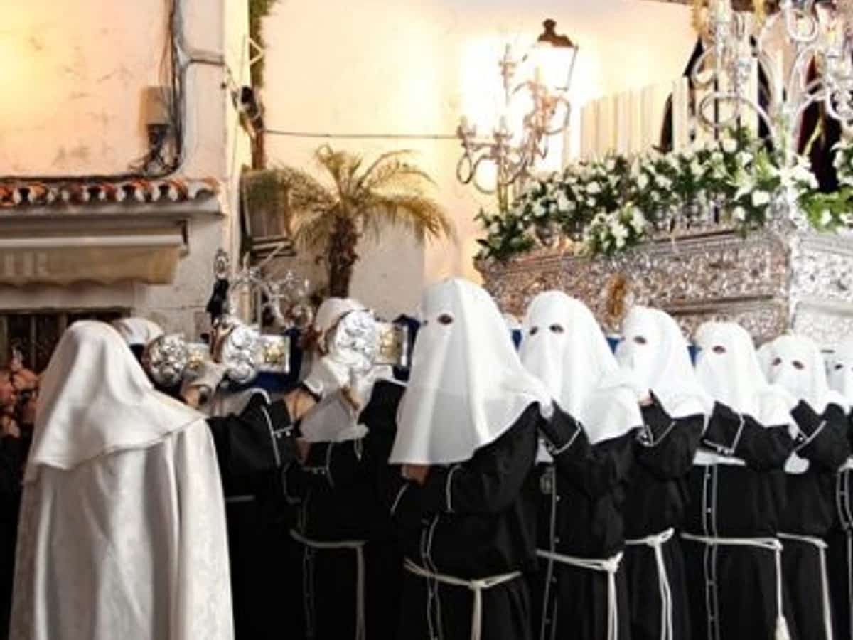 Procesión de Nuestro Padre Jesús Cautivo en Marbella durante el Martes Santo, con nazarenos de túnica negra y capirote blanco acompañando uno de los tronos entre público y balcones llenos en el centro histórico.