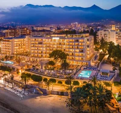 Vista aérea nocturna de un hotel en primera línea de playa en Marbella iluminado, con la ciudad y las montañas al fondo en Navidad Marbella.