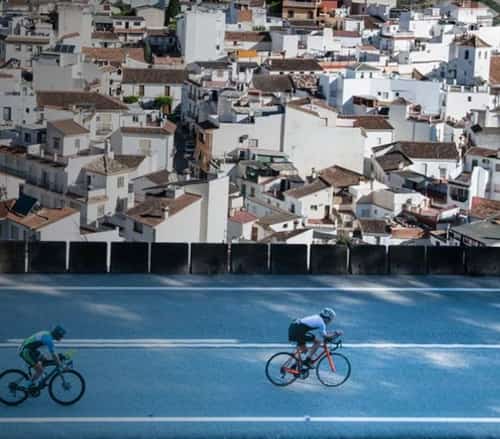 Triatleta corre por la alfombra roja hacia la meta del IRONMAN Marbella mientras el público anima; palmeras y montañas al fondo.