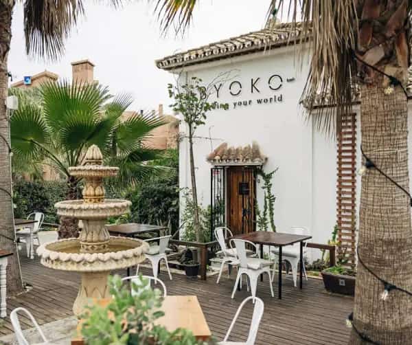 Exterior: White façade of BYOKO Estepona in Urbanización Garden Beach, with stone fountain and wooden terrace surrounded by palm trees and lush Mediterranean greenery.