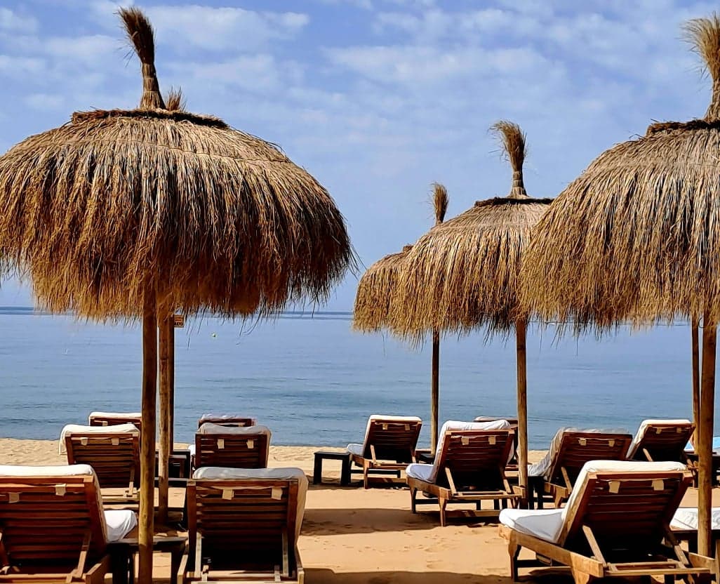 Zona de hamacas con sombrillas de palma en la playa de Río Real, junto a Trocadero Arena Marbella.