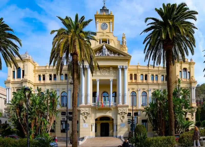 Front façade of Málaga City Hall on Avenida de Cervantes, cream-coloured neo-baroque building with white columns, clock tower and tall palm trees in the foreground on a sunny day.