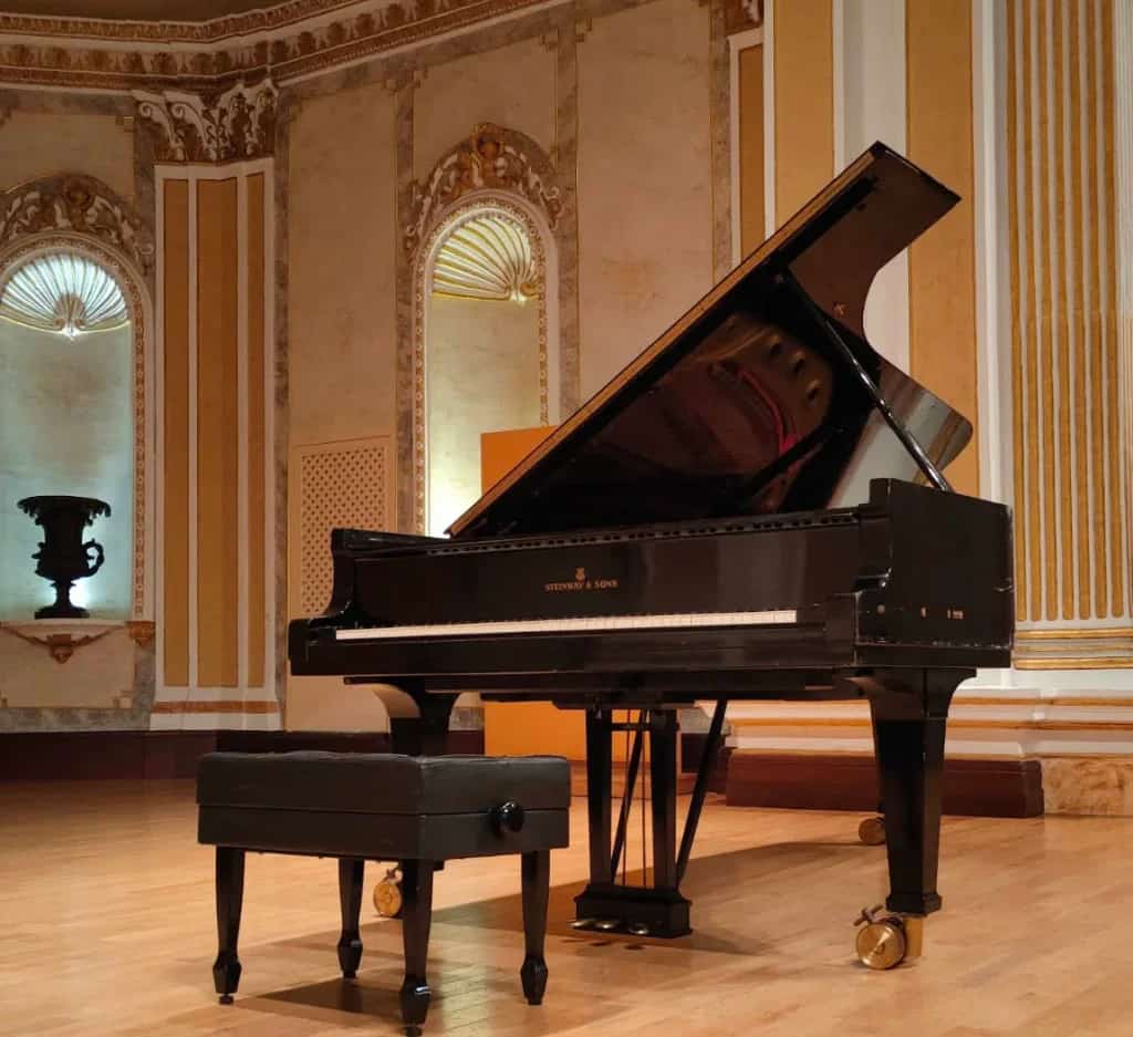 Black Steinway & Sons grand piano on the wooden stage of the María Cristina Hall, with cream and gold decorated walls and lit niches in the background.