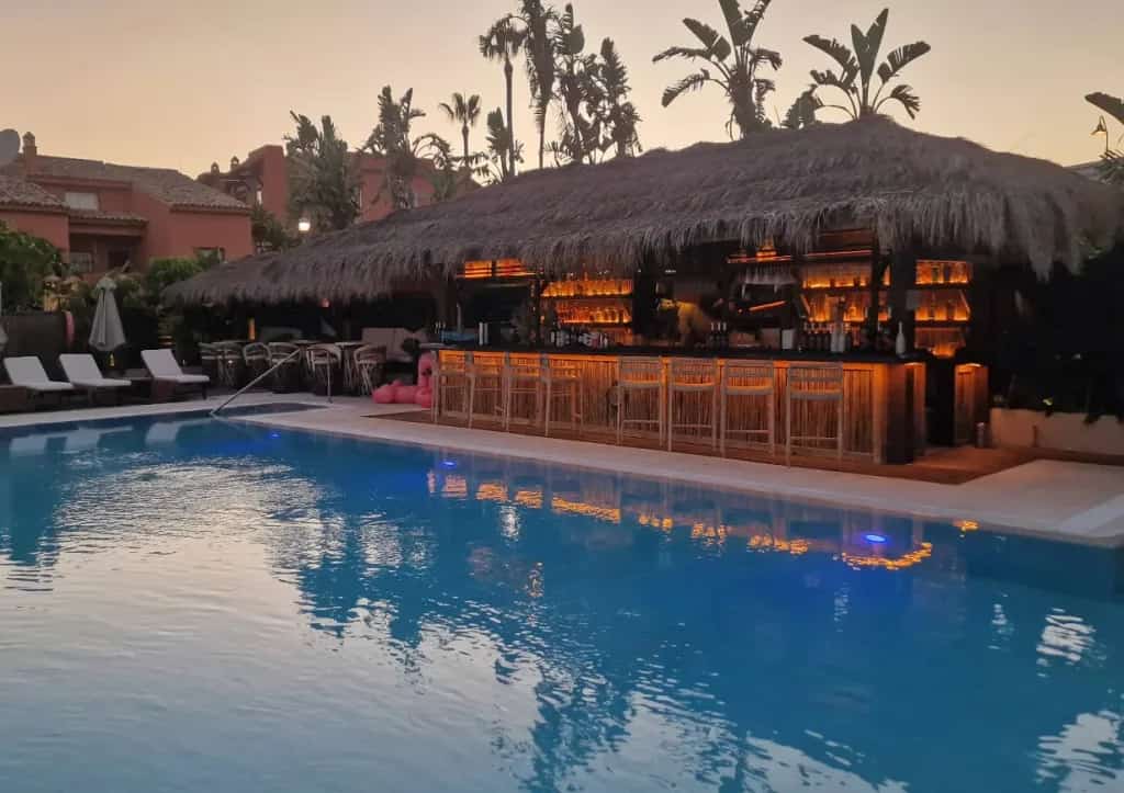 Trocadero Estepona pool bar next to a large swimming pool, with a thatched-roof bar and sun loungers around at sunset.