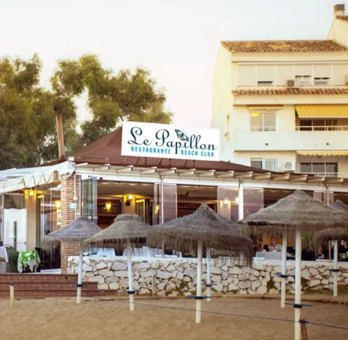 Glass-front dining room at Le Papillon Restaurant & Beach Club overlooking Marbesa Beach and sunbeds.
