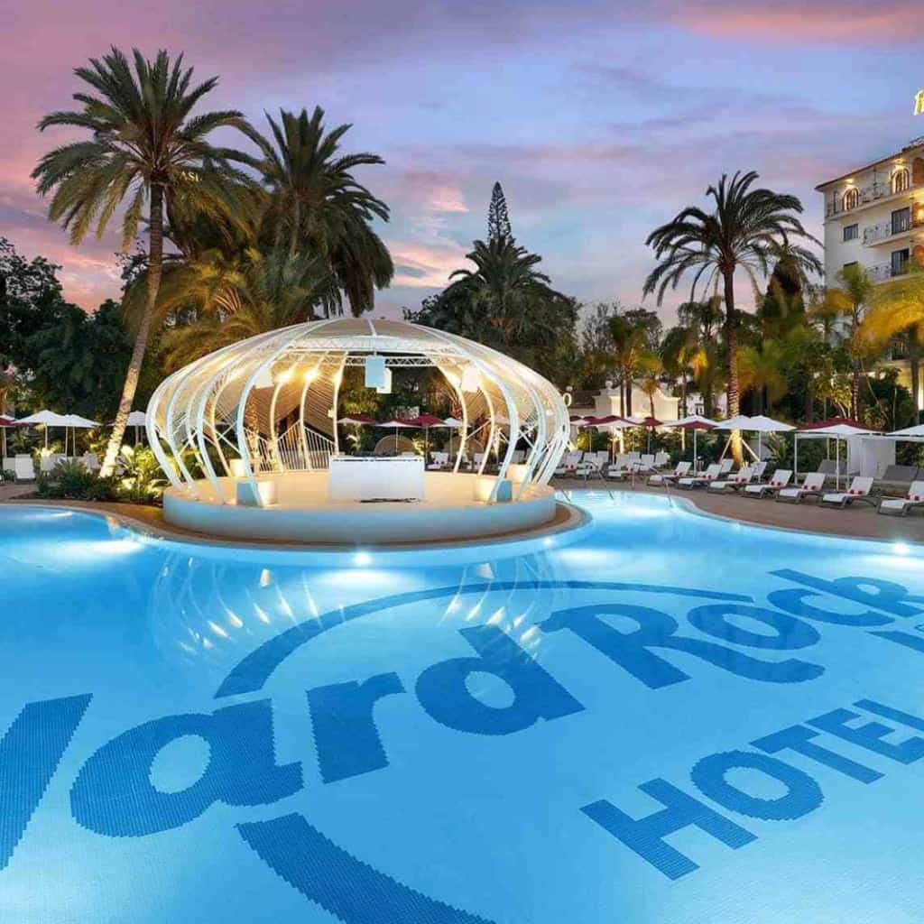 Sunset view of the main pool at Hard Rock Hotel Marbella with the blue hotel logo mosaic on the water and a white illuminated dome structure in the centre, surrounded by sun loungers and palm trees.