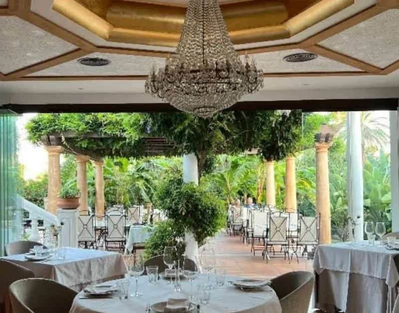 Comedor elegante bajo una lámpara de araña con terraza jardín frondosa en Zeñorío Marbella.
