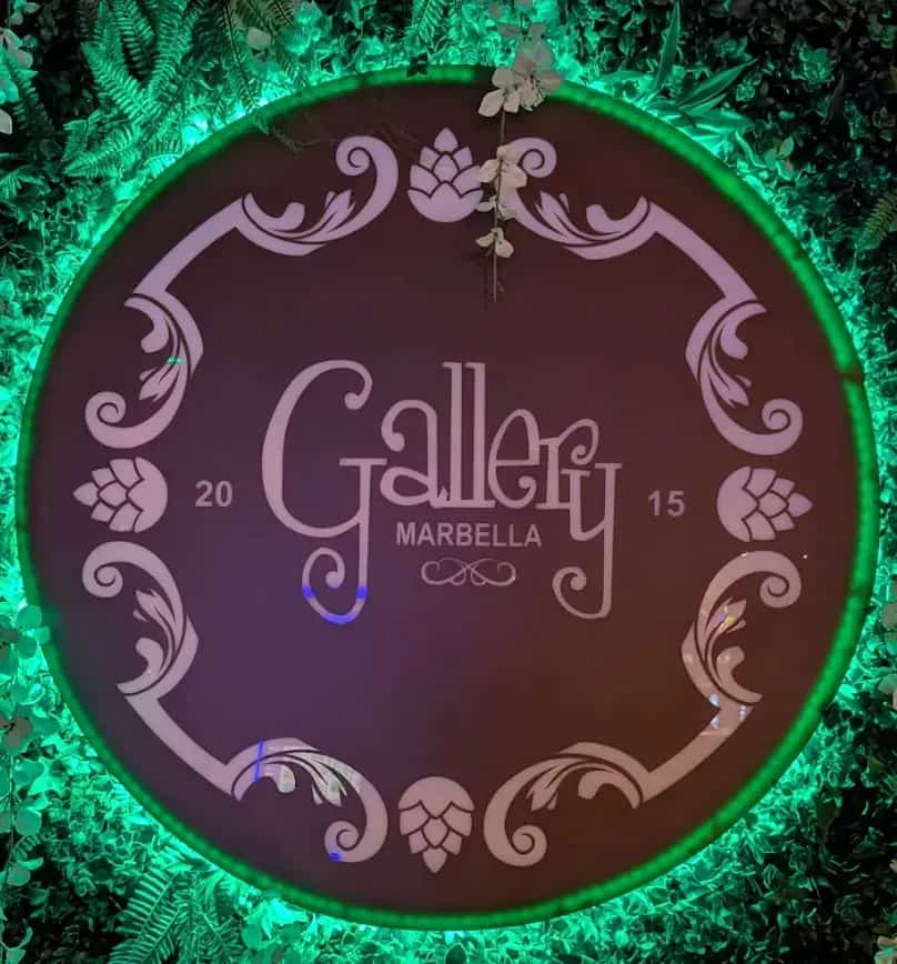 Logo circular iluminado de Gallery Marbella sobre un muro vegetal decorativo con luz verde.